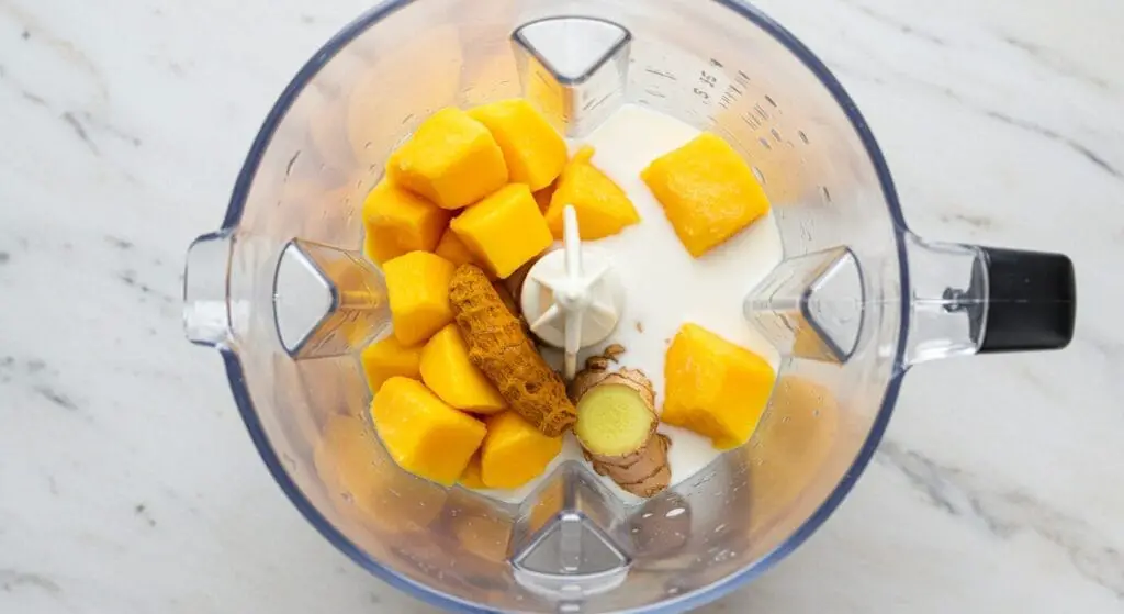 Ingredients for the Turmeric-Mango Smoothie recipe (frozen mango, fresh turmeric root, ginger, plant-based milk, black pepper) being added to a blender pitcher, showcasing the vibrant colors.