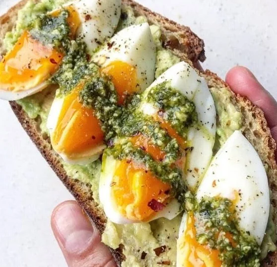 Delicious pesto avocado toast topped with eggs on a wooden table.